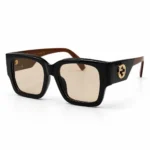 Oversized square sunglasses with a thick glossy black frame and light beige-tinted lenses, shown at a three-quarter angle. The wide temples taper from black to brown, with a gold interlocking-circle logo on the side.