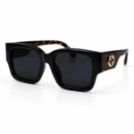 Oversized square sunglasses with a thick glossy black frame and dark gray lenses, shown at a three-quarter angle. The wide temples have a tortoiseshell pattern with a gold interlocking-circle logo on the side.
